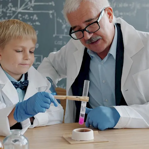 A child in a lab coat and gloves helps an older scientist with test tubes and chemicals in a classroom lab.