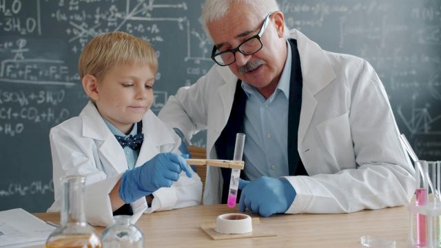 A child in a lab coat and gloves helps an older scientist with test tubes and chemicals in a classroom lab.