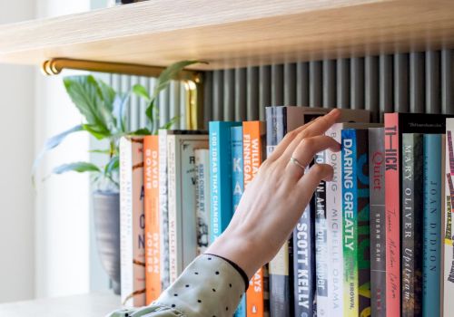 A hand reaches for books on a wooden bookshelf in a bright room.