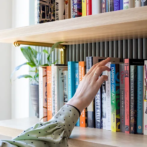 A hand reaches for books on a wooden bookshelf in a bright room.