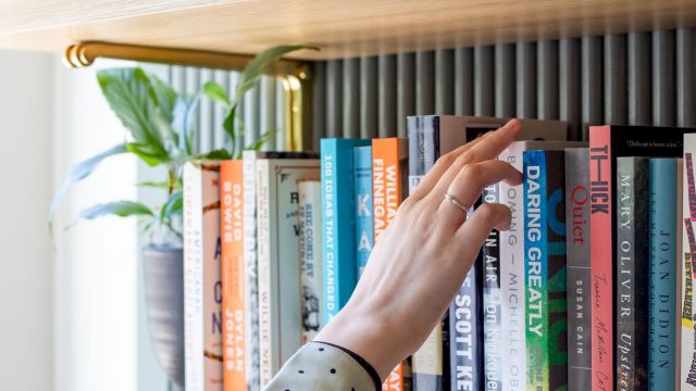 A hand reaches for books on a wooden bookshelf in a bright room.