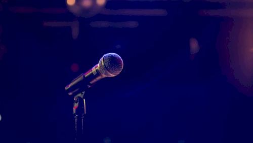 A studio-style microphone on a dark stage under colored lights, with soft bokeh in the background.