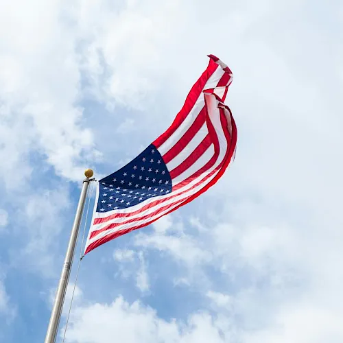A large American flag waves on a pole against a bright blue sky with scattered clouds.