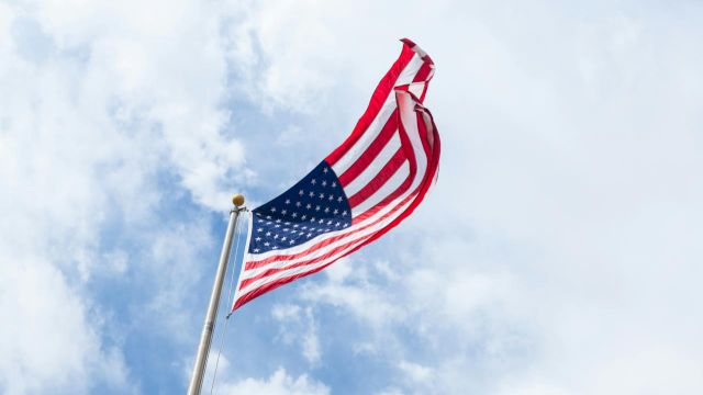 A large American flag waves on a pole against a bright blue sky with scattered clouds.