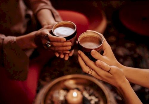 A couple clinks two cups of coffee at a cozy caf&eacute; table in warm golden light, with a reflective tray in view.
