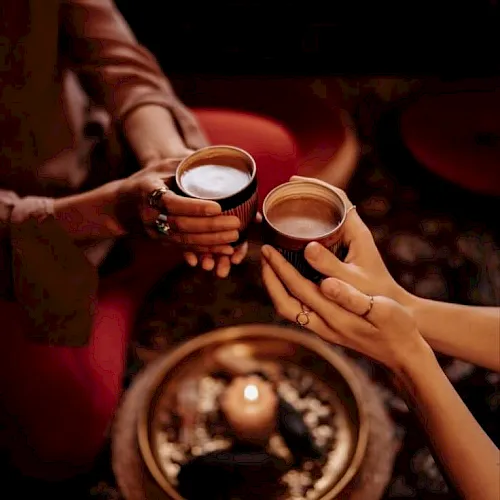 A couple clinks two cups of coffee at a cozy caf&eacute; table in warm golden light, with a reflective tray in view.