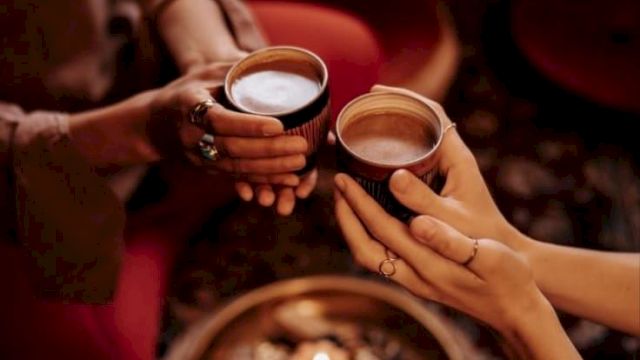 A couple clinks two cups of coffee at a cozy caf&eacute; table in warm golden light, with a reflective tray in view.