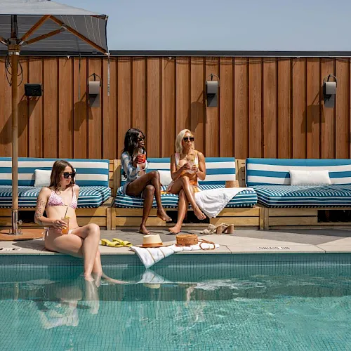 Three women lounge by an outdoor pool under umbrellas on blue-and-white striped cushioned benches, relaxing in the sun.