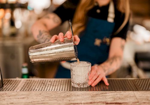 A bartender pours a drink from a metal shaker into a clear glass.