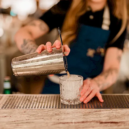 A bartender pours a drink from a metal shaker into a clear glass.