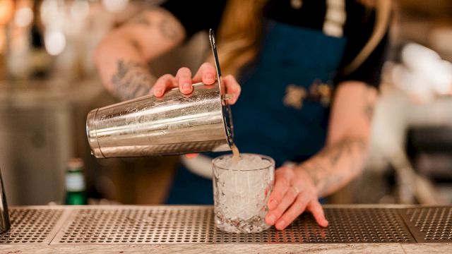 A bartender pours a drink from a metal shaker into a clear glass.