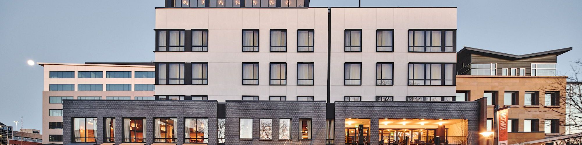 A hotel/office building at dusk with lit windows, a gray facade, and an entrance with outdoor seating.