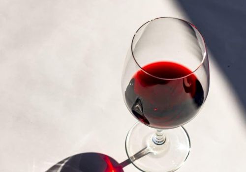 A stemmed glass and a bottle of red wine with sunlight reflections on a white surface.