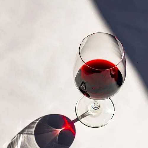 A stemmed glass and a bottle of red wine with sunlight reflections on a white surface.