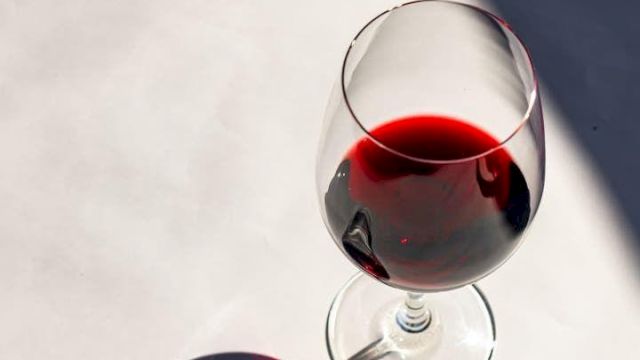 A stemmed glass and a bottle of red wine with sunlight reflections on a white surface.