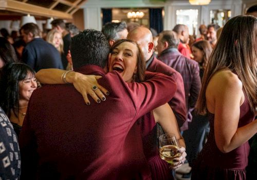 A lively indoor party: a woman in a burgundy dress hugs a man while holding a glass of white wine.