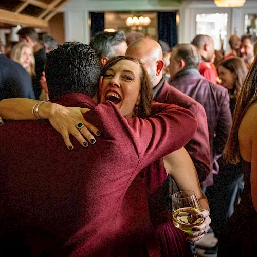 A lively indoor party: a woman in a burgundy dress hugs a man while holding a glass of white wine.