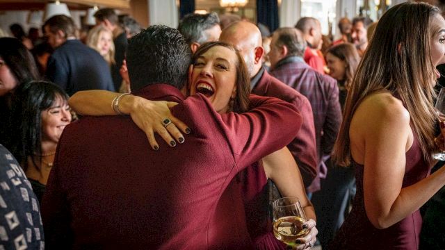 A lively indoor party: a woman in a burgundy dress hugs a man while holding a glass of white wine.