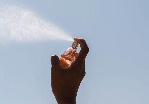 A hand holds a small perfume bottle up against a clear blue sky, catching warm sunlight.