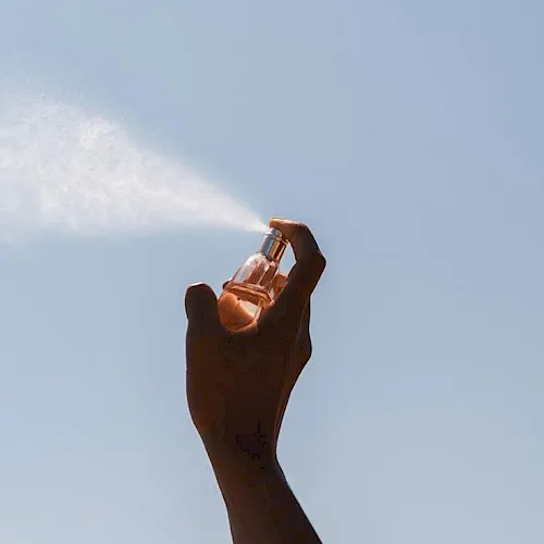 A hand holds a small perfume bottle up against a clear blue sky, catching warm sunlight.
