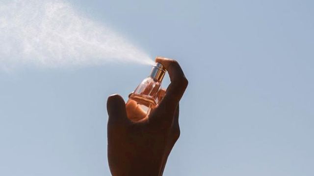 A hand holds a small perfume bottle up against a clear blue sky, catching warm sunlight.