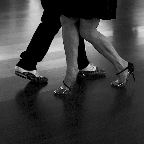 Black-and-white photo of a person&rsquo;s legs walking in shiny high heels and loafers on a tiled floor.