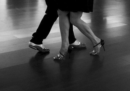 Black-and-white street photo of legs in high heels and flats walking on a tiled floor.