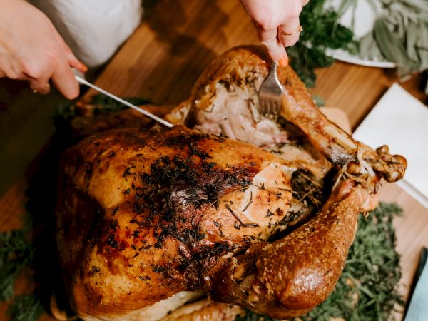 A person carves a whole roast chicken covered in herbs and seasoning outdoors with greenery in the background.