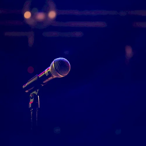 A lit stage microphone on a stand with a blurred performer in the background, captured with colorful concert lighting.