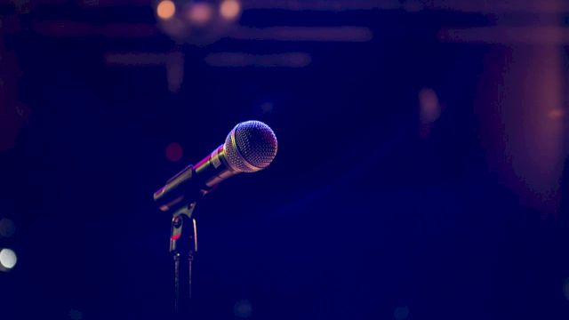 A lit stage microphone on a stand with a blurred performer in the background, captured with colorful concert lighting.