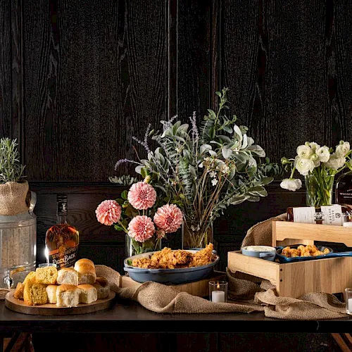 A rustic table set with bouquets of pink/white flowers, candles, bread rolls, and snacks, against dark wooden walls.