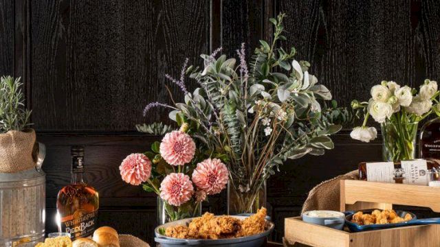 A rustic table set with bouquets of pink/white flowers, candles, bread rolls, and snacks, against dark wooden walls.