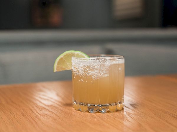 A clear rocks glass holds an iced amber drink, with a lime wedge and sugar-like dust on top.