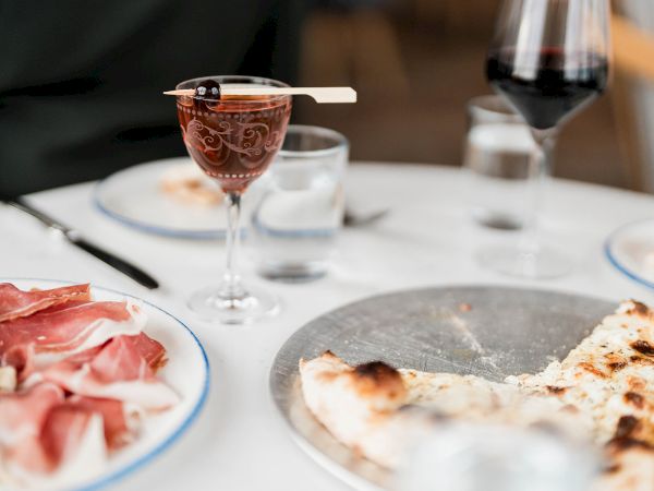 A dinner table with red wine in a goblet, slices of prosciutto, and a partially eaten pizza slice.
