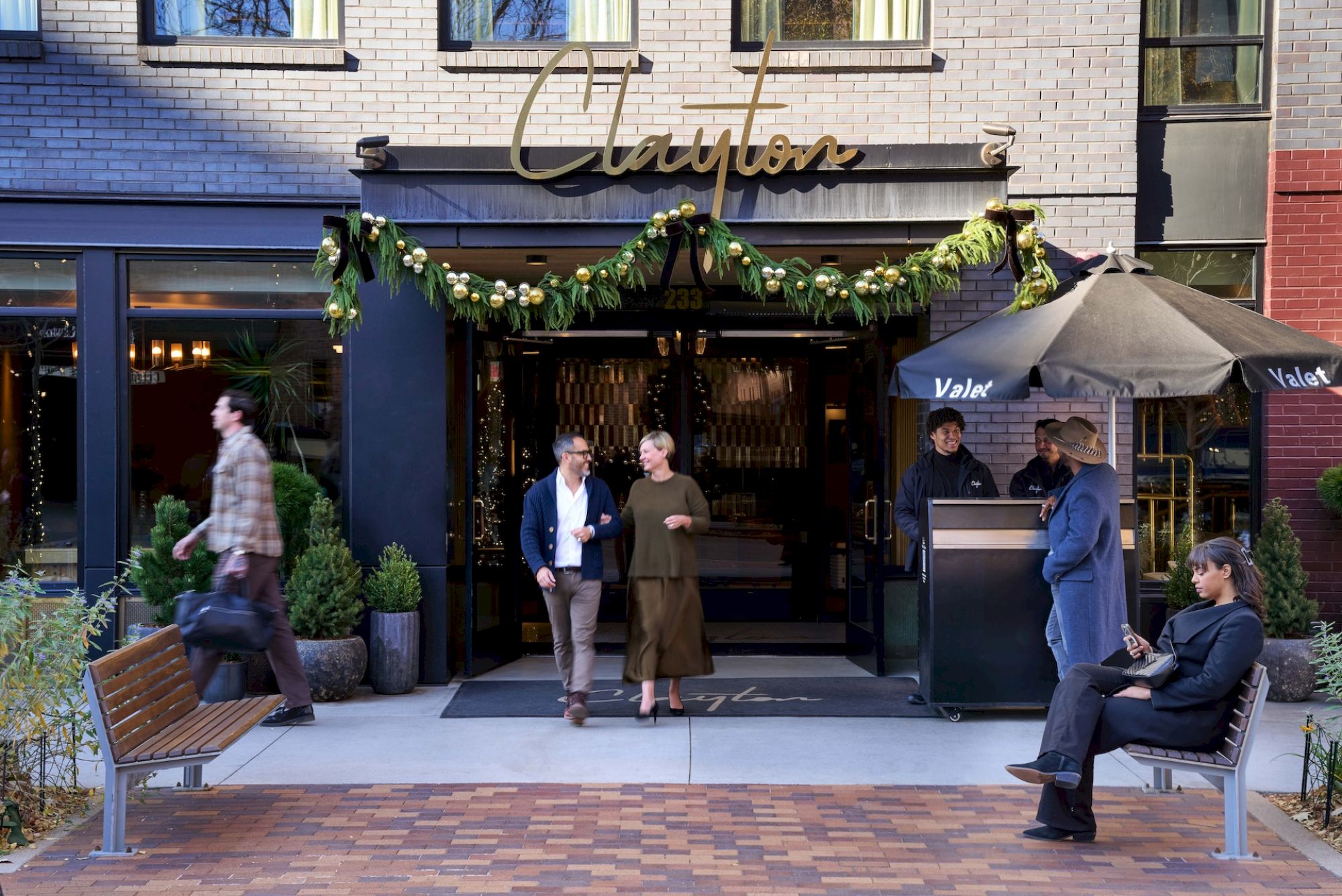 A cozy restaurant entrance with a garlanded awning, people chatting near the doorway, and a few onlookers seated along the brick-paved sidewalk.