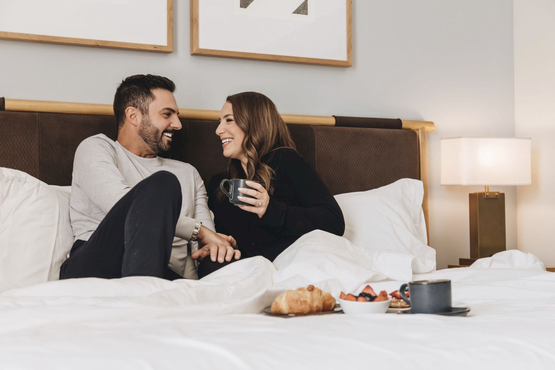 A smiling couple lounges in bed, sharing a warm moment while holding mugs.