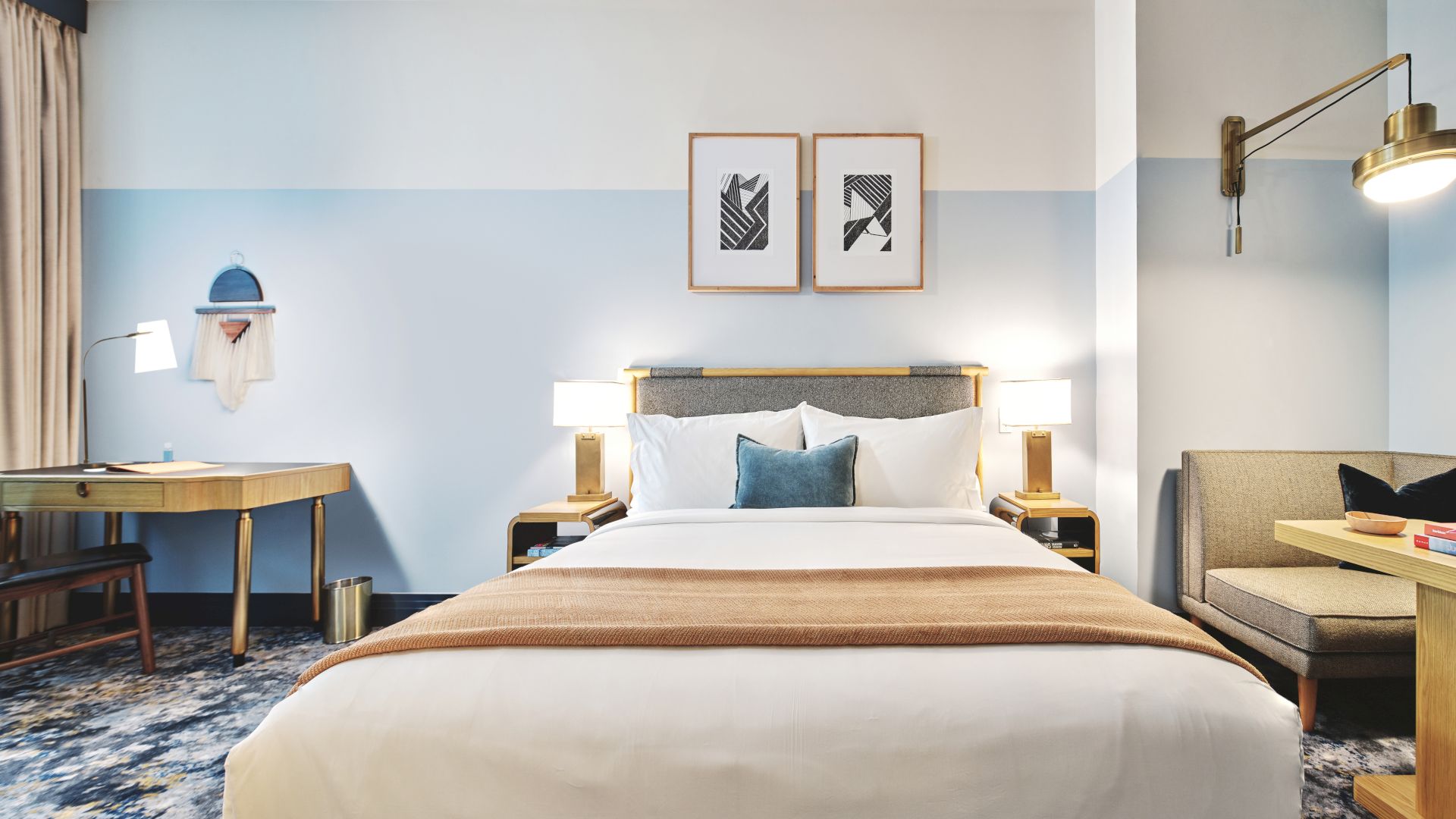 A neatly arranged hotel room features a bed, desk, and seating area with soft lighting and framed art on pastel-colored walls.