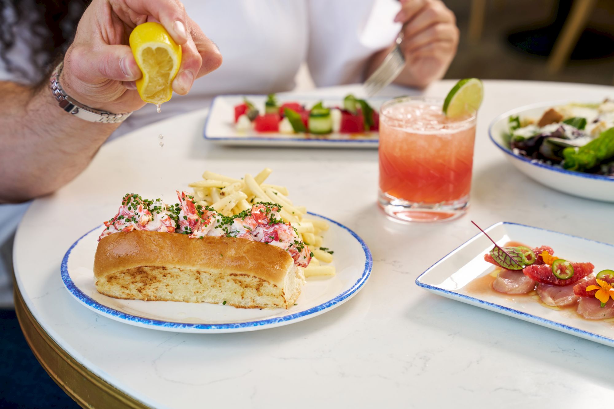 Squeezing lemon over a plate of lobster roll with fries; a pink cocktail and colorful tapas visible in the background, set on a white table.