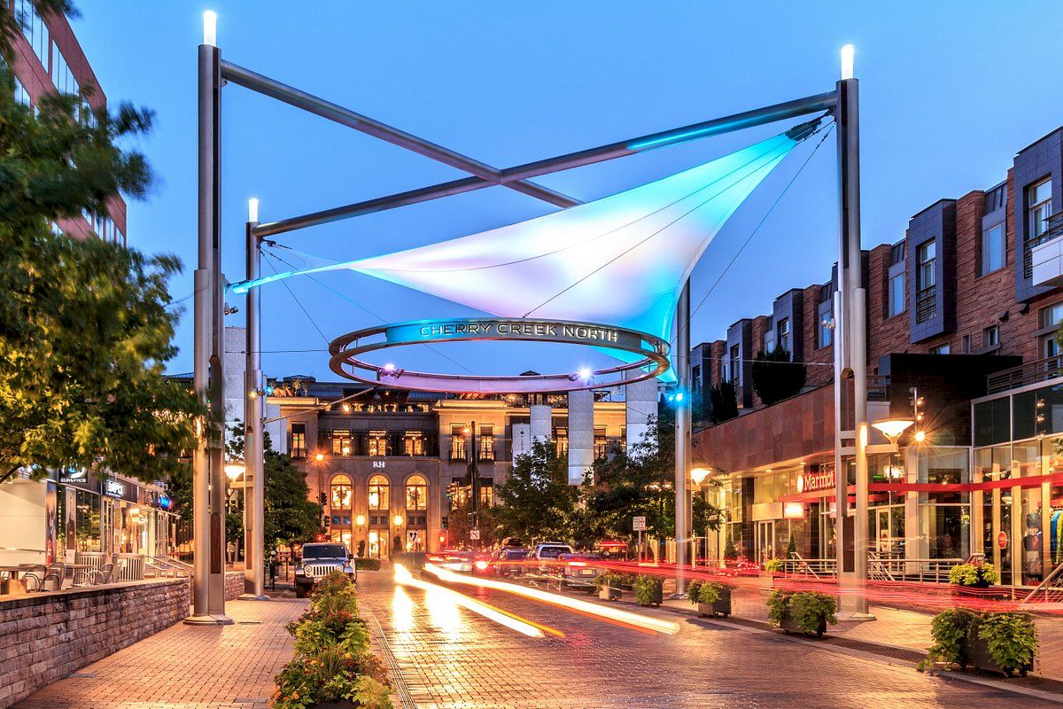A lively urban plaza with modern structures and a circular elevated track, lit by blue lights, at dusk with shops and people around.