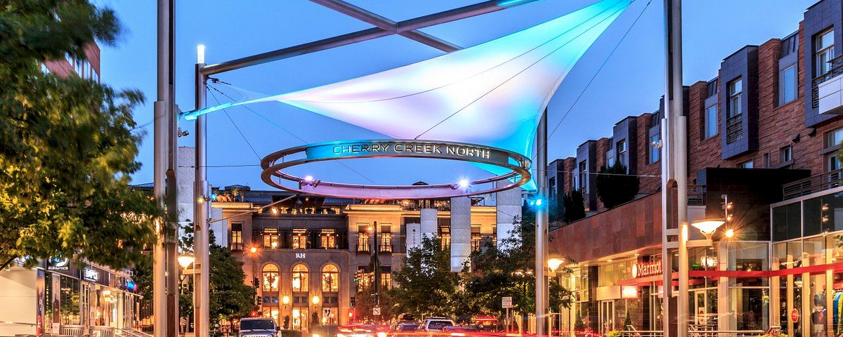 A lively urban plaza with modern structures and a circular elevated track, lit by blue lights, at dusk with shops and people around.