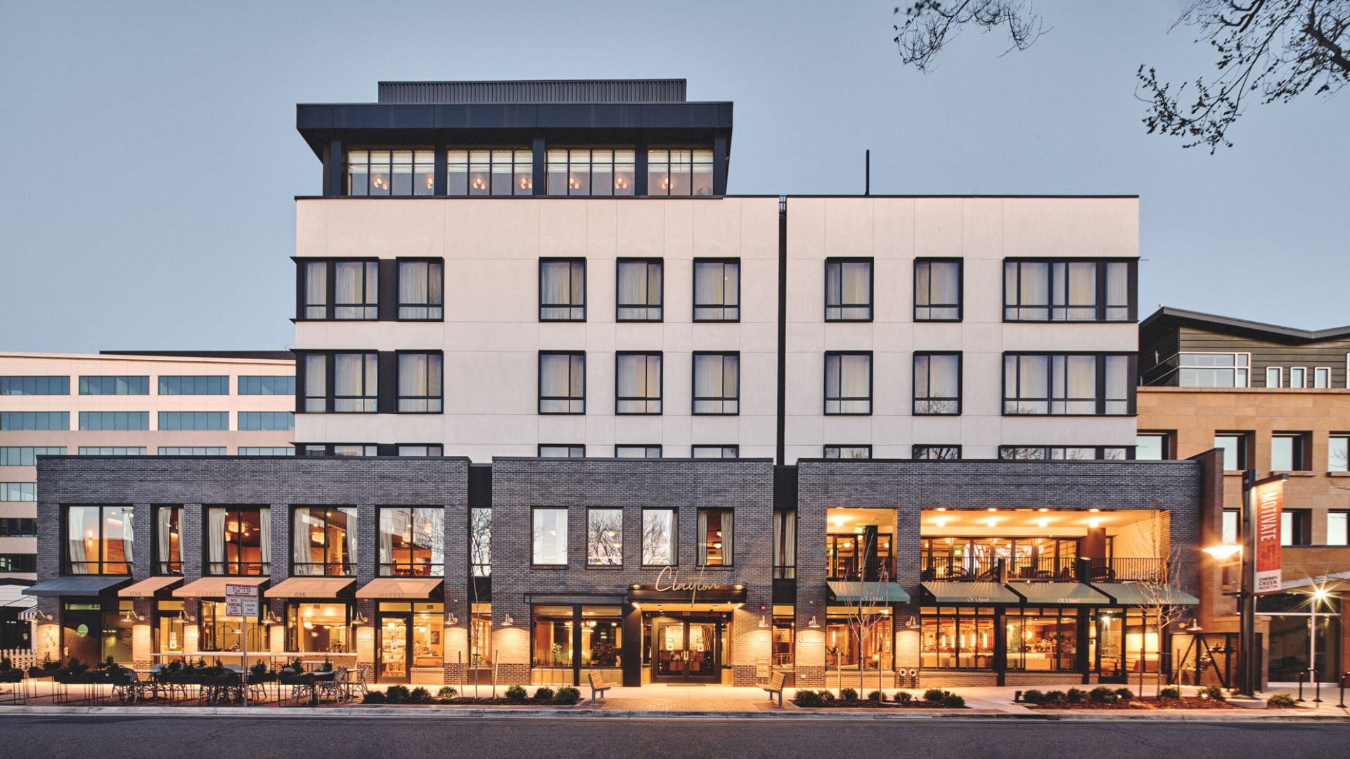 The image shows a modern multi-story building with various shops and businesses on the ground level, featuring large windows and a sleek design.