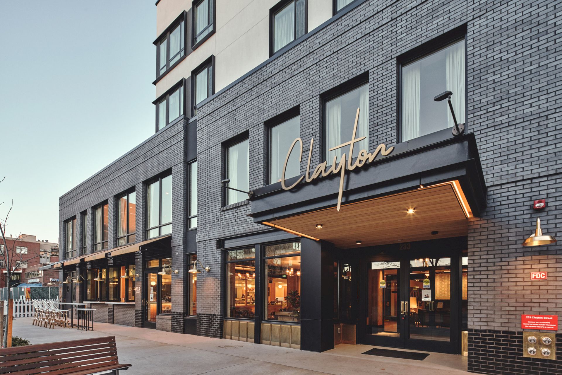 The image shows a modern building with a sign above the entrance that reads "Clayton" in cursive. The facade is made of dark bricks.