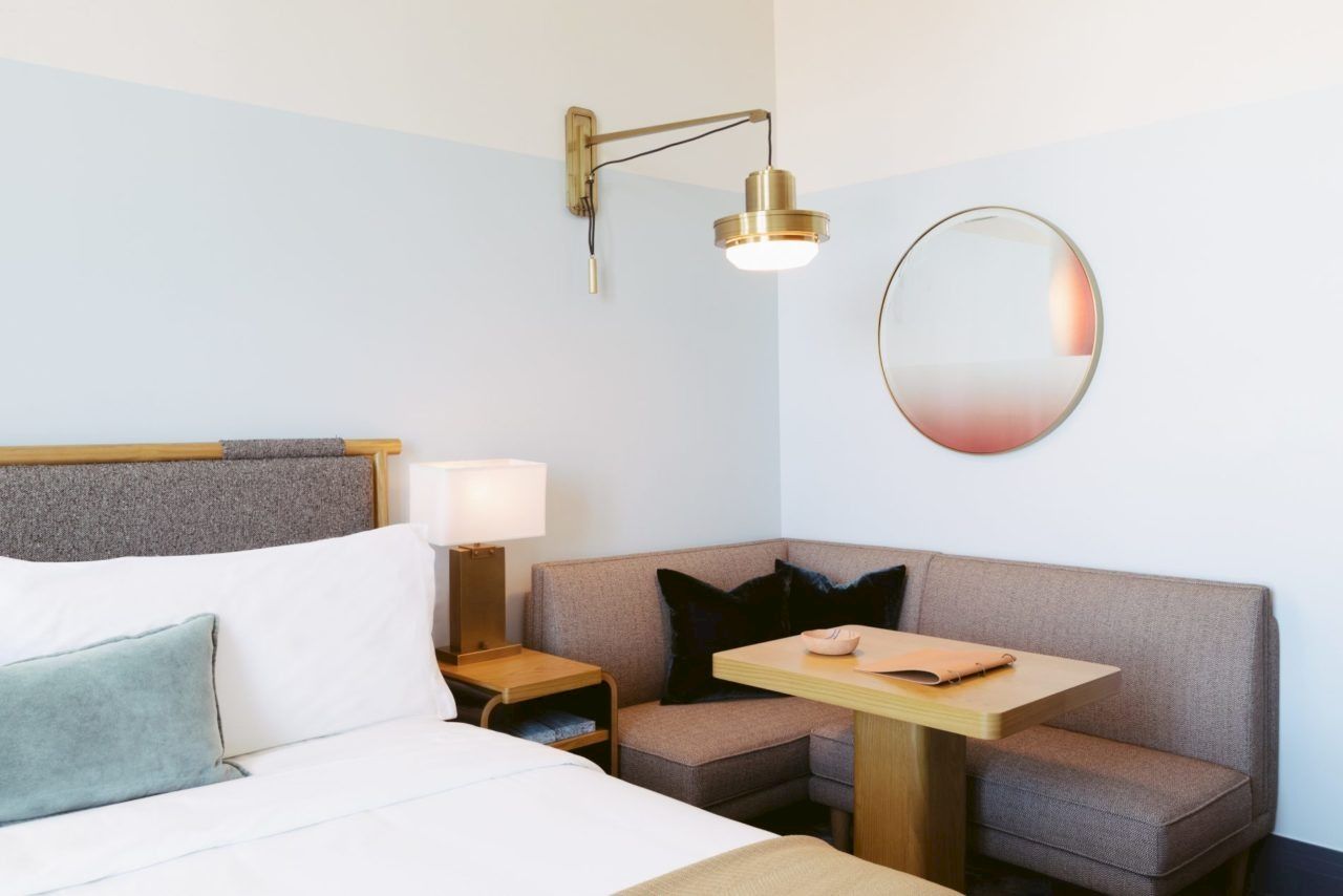 The image shows a modern hotel room with a bed, a lamp, a round wall mirror, and a cozy corner seating area with a small table.