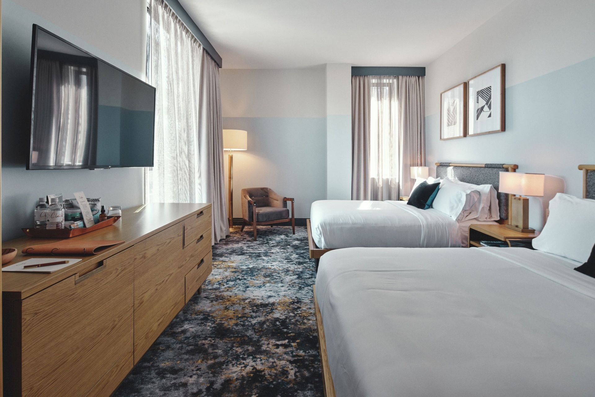 The image shows a hotel room with two beds, a TV, a wooden dresser, a chair, a floor lamp, and wall art, creating a cozy and modern atmosphere.