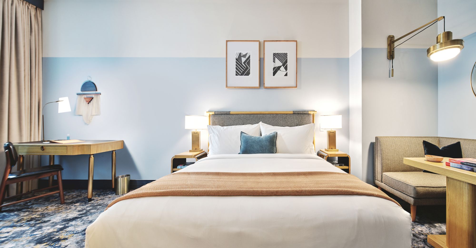 A modern hotel room with a double bed, two side tables, a desk, wall art, and seating area in a light, stylish decor.