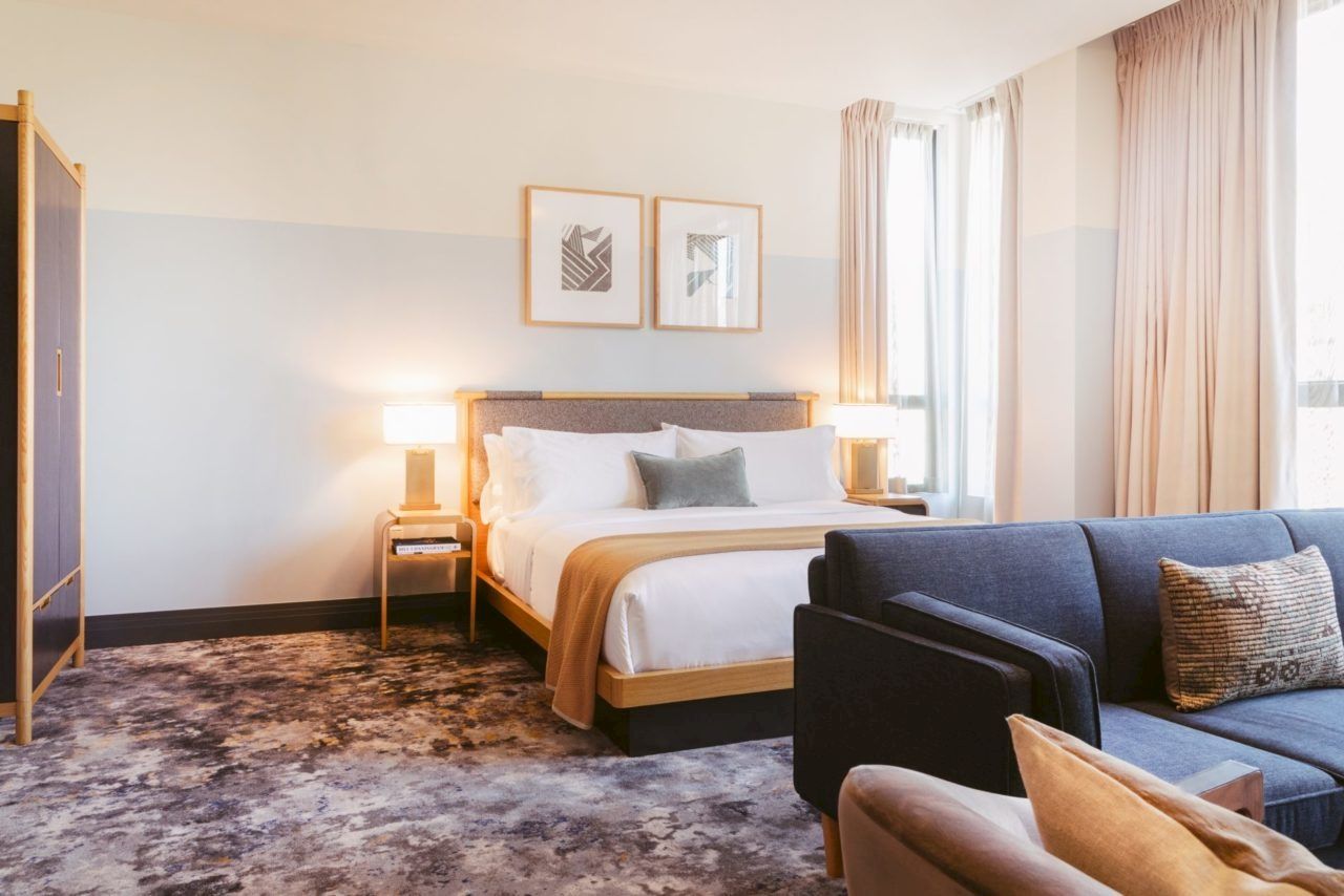 A well-lit hotel room with a bed, two lamps, a sofa, and large windows with curtains, creating a modern and cozy atmosphere.