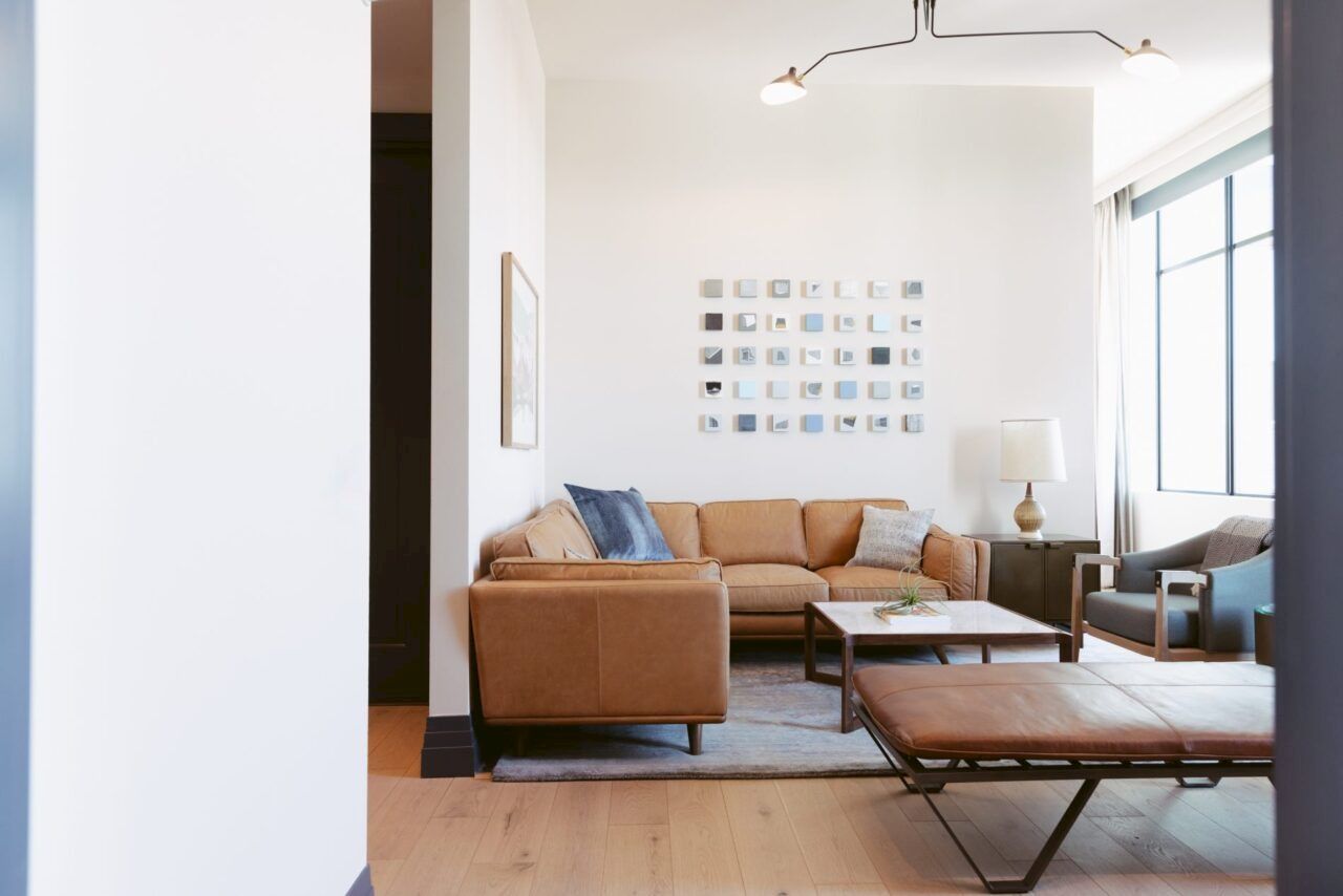 A modern living room features a brown sofa, coffee table, bench, and large window. Wall art and a floor lamp add decor.
