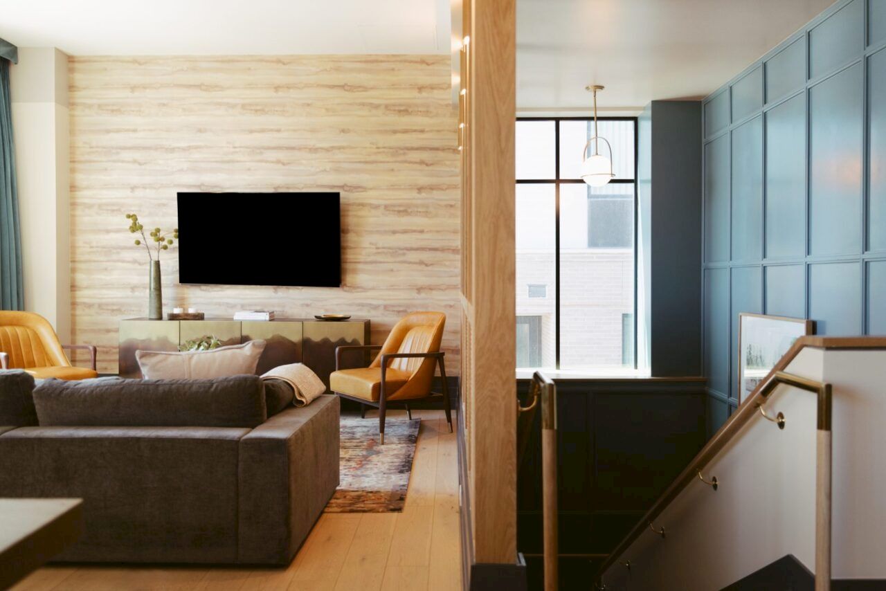 A modern living room with a gray sofa, armchairs, large TV on the wall, and a stairway with blue paneling and a window.