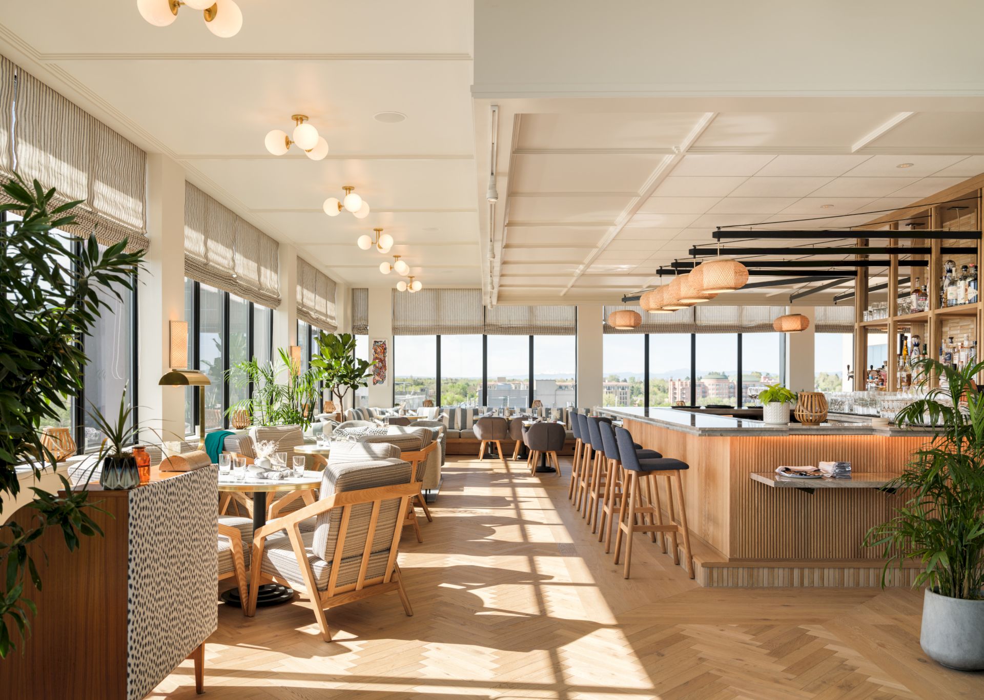 A modern lounge-style restaurant with a long bar, cushioned tables and chairs, pendant lights, large windows, and lots of greenery.