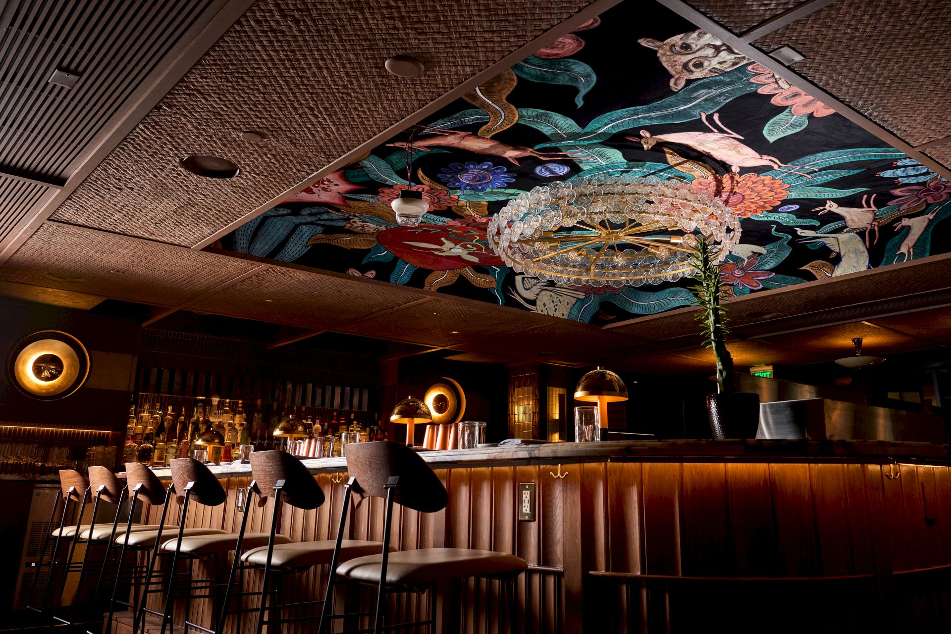 An indoor bar with stools and warm lights, featuring a large colorful ceiling mural above the counter.